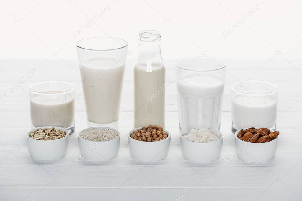 glasses and bottle with coconut, chickpea, oat, rice and almond milk on white wooden table with ingredients in bowls isolated on white