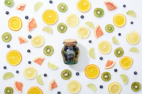 top view of fresh detox drink in jar among sliced fruits and blueberries on white background