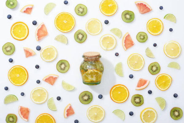 top view of natural detox drink in jar among sliced fruits and blueberries on white background