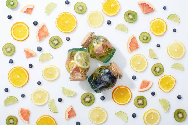 top view of three detox drinks in jars among sliced fruits and blueberries on white background