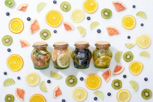 top view of detox drinks in jars among sliced fruits and blueberries on white background