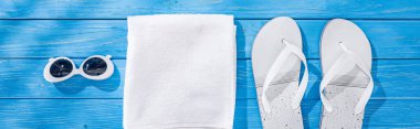 panoramic shot of white folded towel, retro sunglasses and flip flops on blue wooden background clipart
