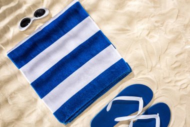 top view of white blue striped folded towel, retro sunglasses and flip flops on sand  clipart
