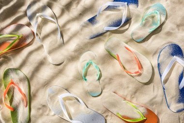 top view of scattered turquoise, orange, blue and green flip flops on sand with shadows clipart