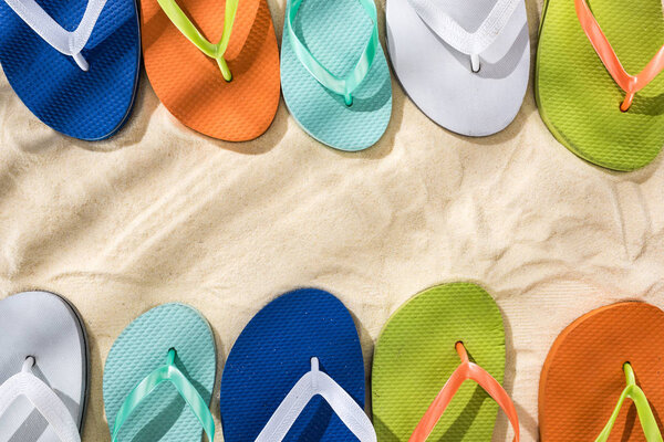 top view of white, green, orange, turquoise and blue flip flops on sand 