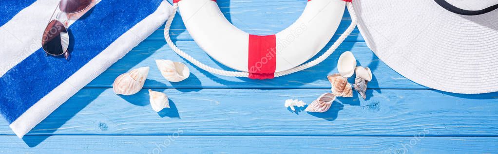panoramic shot of striped towel, sunglasses, lifebuoy, white floppy hat and seashells on blue wooden background