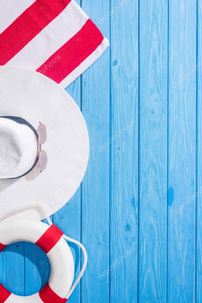 top view of striped towel, white sunglasses, lifebuoy, floppy hat on blue wooden background with copy space