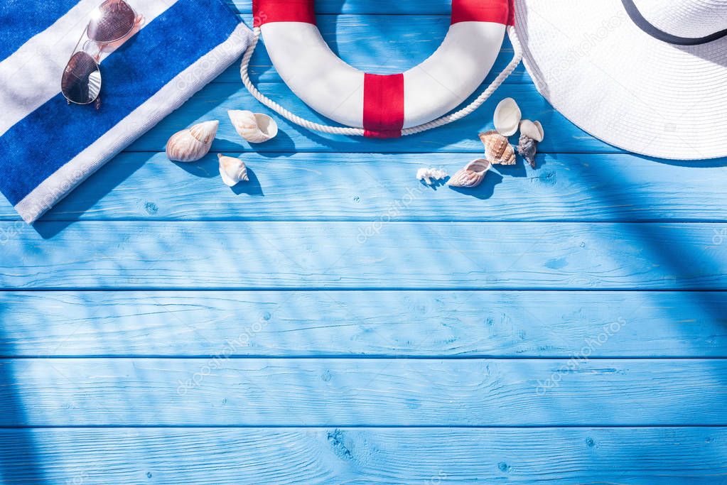 top view of striped towel, sunglasses, lifebuoy, white floppy hat and seashells on blue wooden background with shadows and copy space