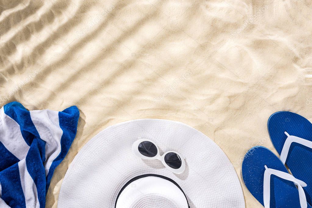 top view of striped towel, retro sunglasses, floppy hat and blue flip flops on sand with shadows and copy space