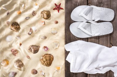 top view of seashells and starfish on sand and flip flops and white towel on wooden brown board clipart