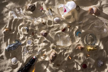 deniz kabukları, cam şişe, dağınık sigara izmaritleri, kırık bardak, elma çekirdeği, plastik bardak ve gölgeler ile kum üzerinde şeker sarıcı üst görünümü