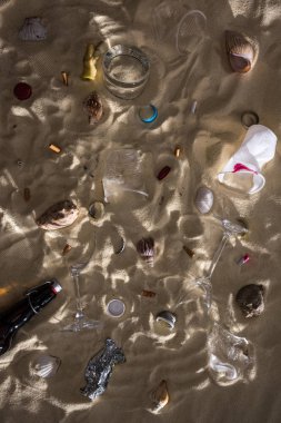 top view of seashells, glass bottle, scattered cigarette butts, broken glasses, apple core, plastic cups and candy wrapper on sand with shadows