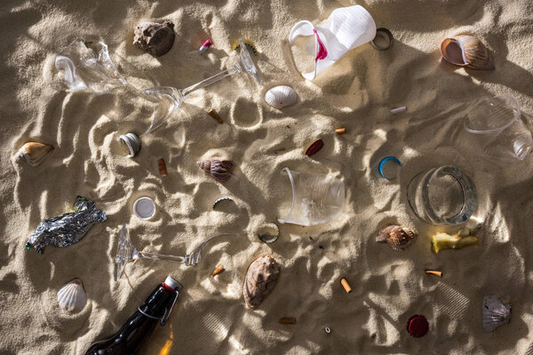 вид сверху ракушек, стеклянная бутылка, разбросанные окурки, разбитые бокалы, яблочная сердцевина, пластиковые чашки и обертка от конфет на песке с тенями
