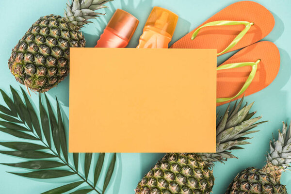 top view of pineapples, tropical leaf, sunscreens, orange flip flops and empty card on turquoise background