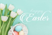 top view of painted chicken eggs in wicker basket and white tulips on blue background with white happy Easter lettering 