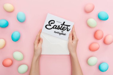 cropped view of woman holding envelope and greeting card with happy Easter to everyone lettering near multicolored painted chicken eggs on pink background  clipart