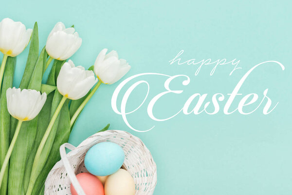 top view of painted chicken eggs in wicker basket and white tulips on blue background with white happy Easter lettering 