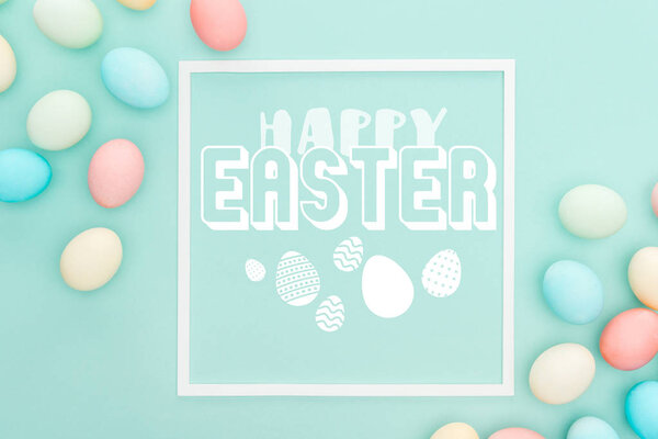 top view of happy Easter lettering in white square frame near painted chicken eggs on blue background