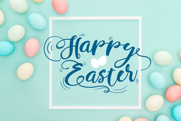 top view of happy Easter blue lettering in white square frame near painted chicken eggs on blue background
