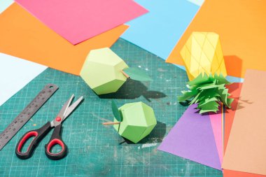 origami fruits and colorful cardboard with scissors and ruler on messy surface clipart