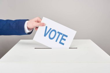 partial view of man putting card with vote in box on grey background clipart