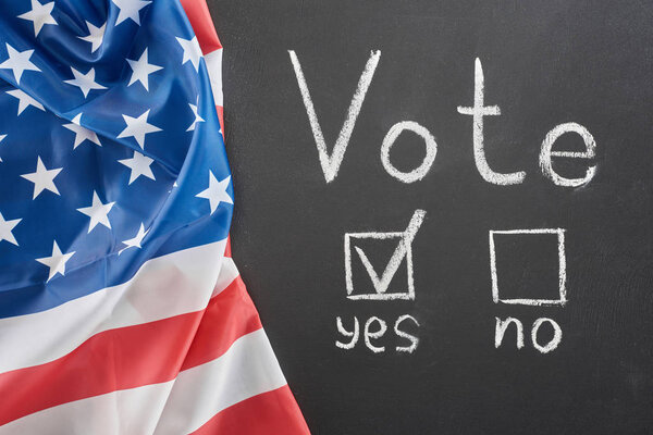 Top view of white vote lettering and check mark near yes word on black chalkboard near american flag
