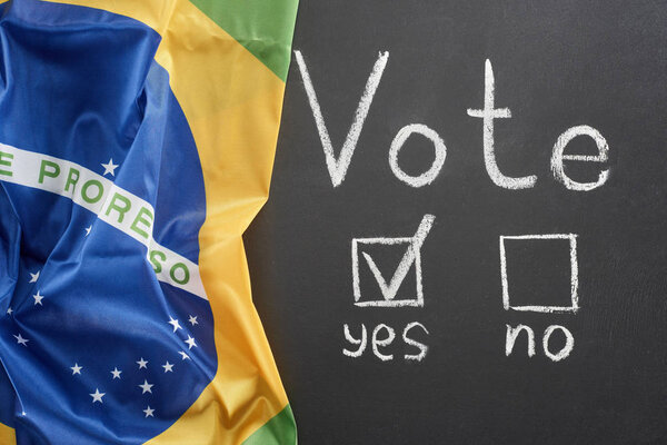 Top view of white vote word and check mark near yes word on black chalkboard near flag of Brazil

