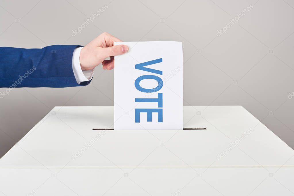 cropped view of man putting card with vote in box on grey background