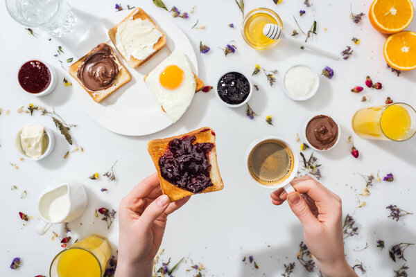 cropped view of woman holding cup with coffee and toast with jam on white 