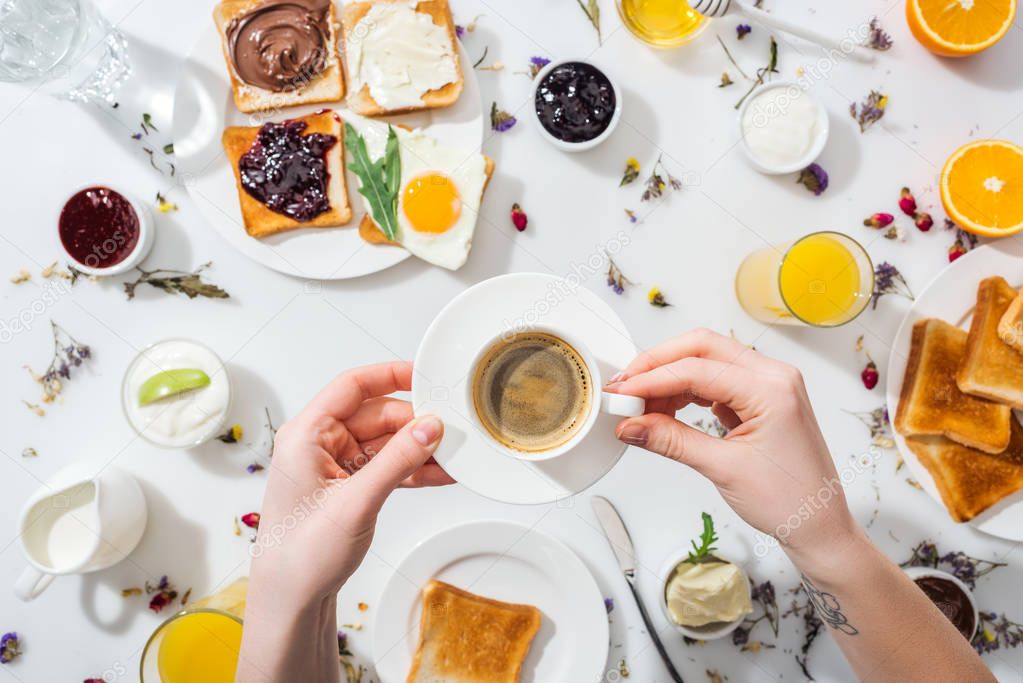 cropped view of woman with tattoo on hand holding saucer and cup of espresso near breakfast on white 