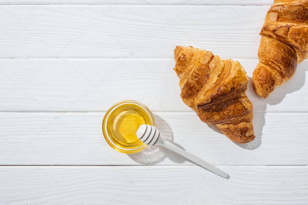 top view of tasty and fresh croissants near glass bowl with honey and honey dipper on white surface 