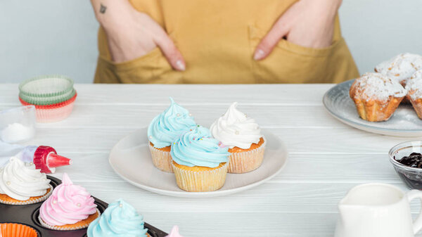 Cropped view of woman with hands in pockets near table with cupcakes isolated on grey