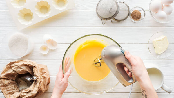 Cropped view of woman mixing dough with hand mixer on wooden table with ingredients