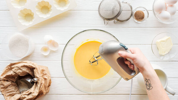 Cropped view of woman mixing dough with hand mixer on wooden table with ingredients