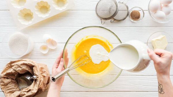 Cropped view of woman pouring milk in liquid dough on table with ingredients