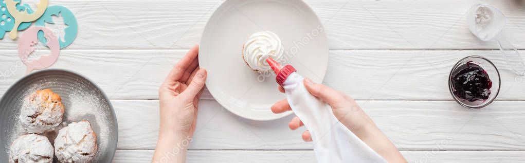 panoramic shot of woman decorating cupcake with icing bag on white wooden table
