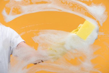 cropped view of cleaner in rubber glove cleaning glass with sponge on orange background clipart