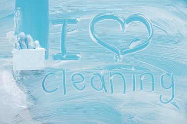 partial view of cleaner with sponge near i love cleaning handwritten lettering on glass with white foam on blue background clipart
