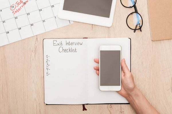 cropped view of recruiter holding smartphone with blank screen above notebook with exit interview checklist lettering