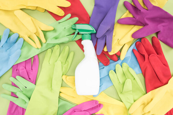 top view of messy scattered multicolored rubber gloves and white spray bottle with cleanser on green background