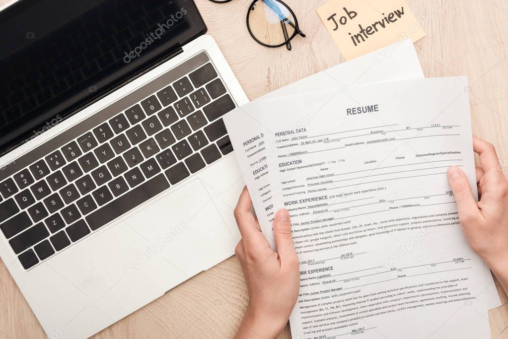 cropped view of recruiter holding resume templates near sticky note with job interview lettering and laptop