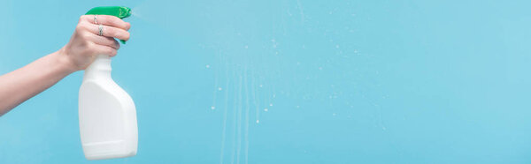 panoramic shot of woman spraying glass with detergent on blue background