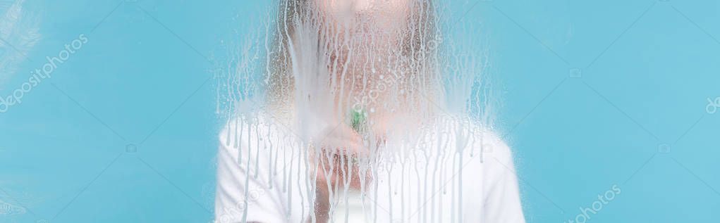 panoramic shot of young woman cleaning glass with dripping detergent on blue background