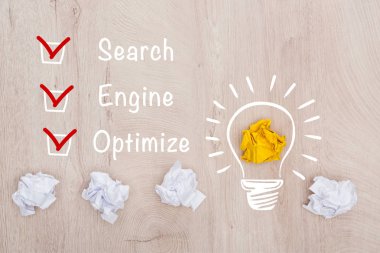 Arama, motor yakınlarındaki buruşuk kağıt topların üst görünümü, kelimeleri optimize et ve ahşap yüzey üzerindeki ampul illüstrasyonu, iş konsepti