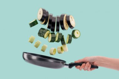 Cropped view of woman holding frying pan with sliced fresh vegetables above isolated on turquoise  clipart