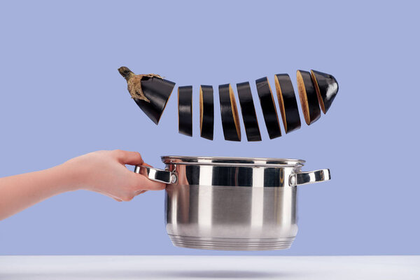 Cropped view of woman holding stainless steel saucepan with chopped raw organic aubergine above white surface isolated on blue