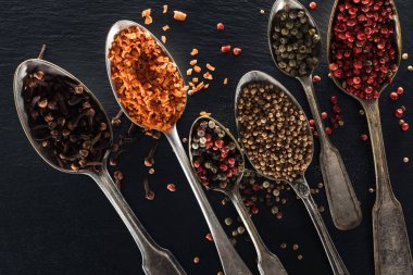 top view of pepper, candied fruit, coriander and carnation seeds in silver spoons on black textured background clipart
