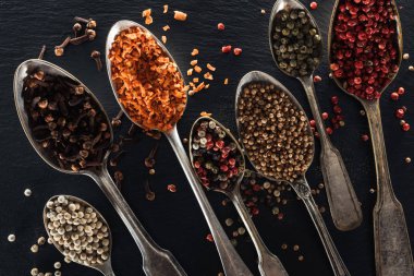 top view of indian spices in silver spoons on black textured background clipart