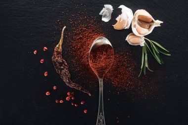 top view of red pepper powder in silver spoon on black background with dried chili peppe, herbs and garlic clipart