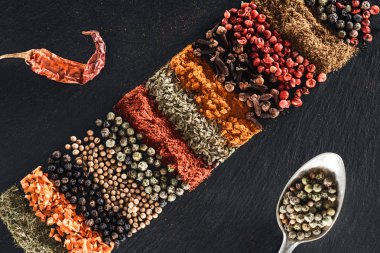 top view of traditional indian spices on textured black background near spoon with white pepper and dried chili pepper clipart